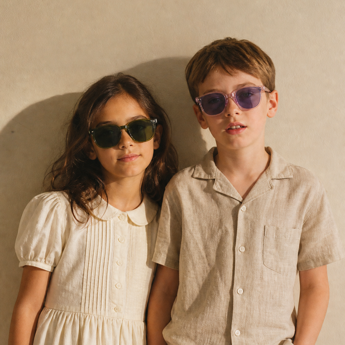 Two children standing against a textured beige wall, wearing casual light-colored clothing and sunglasses. The child on the left wears a cream puff-sleeve dress, and the child on the right wears a beige short-sleeve button-up shirt with white pants.